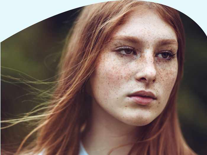 Ein Porträt einer jungen Frau mit langen, roten Haaren und Sommersprossen. Sie schaut seitlich weg mit einem neutralen Gesichtsausdruck. Der Hintergrund ist unscharf, mit dem Anschein von natürlicher Vegetation.